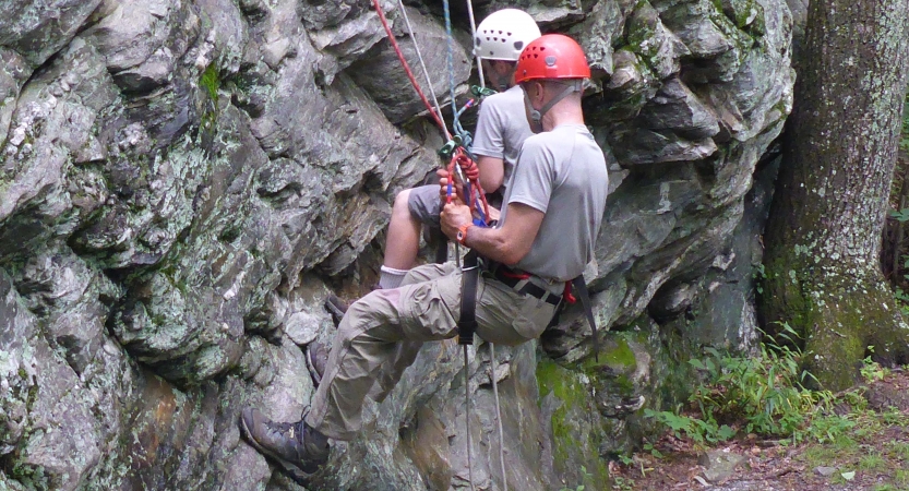 Two people wearing safety gear are secured by ropes as they sit back into their harnesses while taking a break from rock climbing.